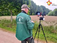 Ein Drohnenpilot mit der Rückenaufschrift "Kitzrettung Hegering Rietberg" auf der grünen Jacke steht hinter einer auf einem Stativ montierten Kamera.