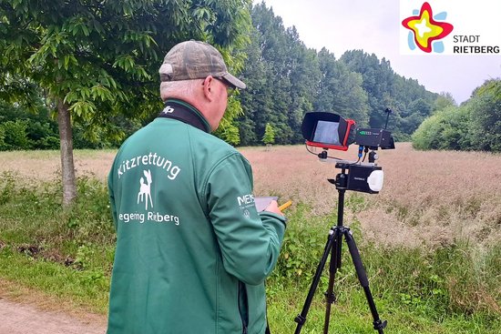 Ein Drohnenpilot mit der Rückenaufschrift "Kitzrettung Hegering Rietberg" auf der grünen Jacke steht hinter einer auf einem Stativ montierten Kamera.