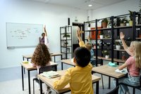 Mehrere Kinder in einer Schulklasse sind von hinten an ihren Tischen sitzend zu sehen, während eine Lehrerin vorn an der Tafel stehend unterrichtet.