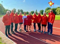 Mehrere Männer und Frauen stehen in rote Jacken gewandet auf einem Sportplatz. Die Dame in weißer Bluse in der Mitte hält eine der roten Jacken mit der Rückseite zur Kamera.