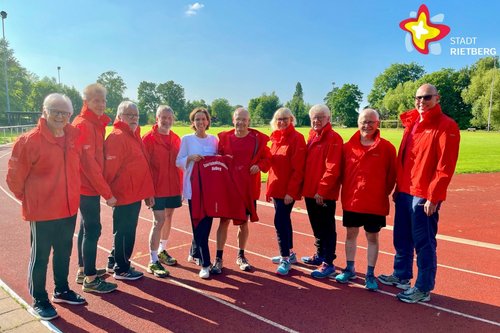 Mehrere Männer und Frauen stehen in rote Jacken gewandet auf einem Sportplatz. Die Dame in weißer Bluse in der Mitte hält eine der roten Jacken mit der Rückseite zur Kamera.