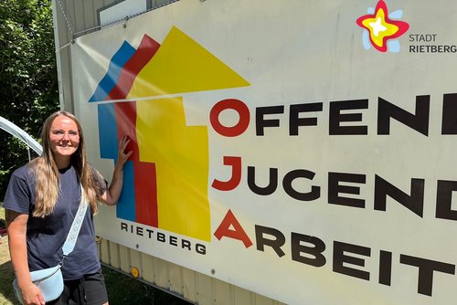 Julia Schmitfranz steht draußen an eine Wand gelehnt, die die Aufschrift Offene Jugend Arbeit trägt.