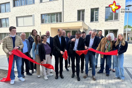 Etwa 25 Personen stehen hinter einem breiten roten Band. Drei Personen schneiden das Band in der Mitte durch. Im Hintergrund ist das Schulgebäude zu sehen.