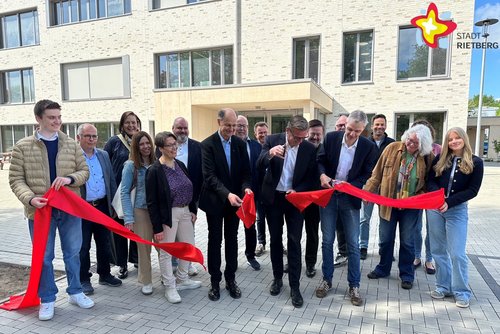 Etwa 25 Personen stehen hinter einem breiten roten Band. Drei Personen schneiden das Band in der Mitte durch. Im Hintergrund ist das Schulgebäude zu sehen.