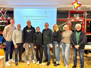 Fünf Männer und drei Frauen stehen lächelnd nebeneinander. Im Hintergrund ist auf einer Leinwand der Schriftzug "Mitgliederversammlung Stadtsportverband" zu lesen.