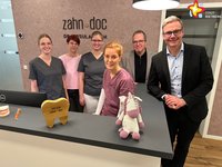 Die genannten Personen stehen hinter dem Empfangstresen der Praxis und schauen in die Kamera.