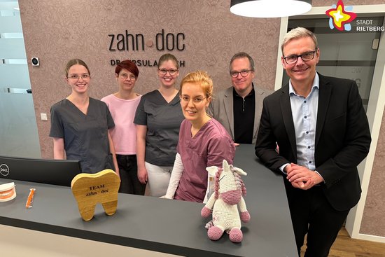 Die genannten Personen stehen hinter dem Empfangstresen der Praxis und schauen in die Kamera.