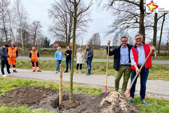 Ortsvorsteher Detlev Hanemann und Bürgermeister Andreas Sunder stehe mit Schaufeln in der Hand neben einer jungen Linde, die soeben in ein noch offenes Pflanzloch gesetz wurde. Im Hintergrund, am Rande eines radweges, stehen Christian Schlingschrörder, Petra und Marvin Busche, Anja Rodenbeck, Alexander Speter, Martin Böcker und Tobias Keßler. Foto Stadt Rietberg