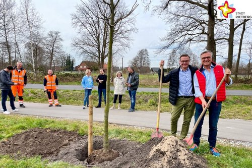 Ortsvorsteher Detlev Hanemann und Bürgermeister Andreas Sunder stehe mit Schaufeln in der Hand neben einer jungen Linde, die soeben in ein noch offenes Pflanzloch gesetz wurde. Im Hintergrund, am Rande eines radweges, stehen Christian Schlingschrörder, Petra und Marvin Busche, Anja Rodenbeck, Alexander Speter, Martin Böcker und Tobias Keßler. Foto Stadt Rietberg