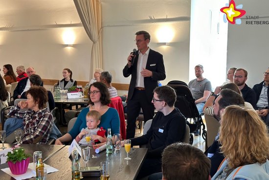Die Gäste sitzen an Tischen im Ratssaal und hören Bürgermeister Andreas Sunder zu. Er steht in der Mitte und hält ein Mikrofon in der Hand.