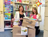 Zwei Frauen knien vor einem Bücherregal und hinter vier Kartons und halten je ein Bilderbuch in der Hand.