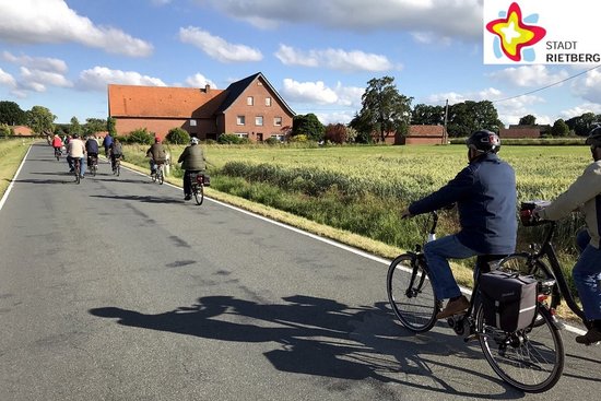 Unter mit Wolken bedecktem blauen Himmel fahren mehrere Radfahrer hintereinander auf einer aspahltierten Straße durch die nur mäßig besiedelte Landschaft.