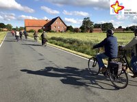 Unter mit Wolken bedecktem blauen Himmel fahren mehrere Radfahrer hintereinander auf einer aspahltierten Straße durch die nur mäßig besiedelte Landschaft.