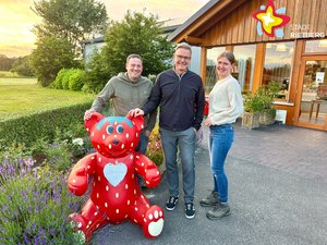 Zwei Männer und eine Frau stehen hinter einer als Erdbeere lackierten Bären-Figur und lachen in die Kamera.