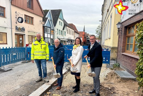 Drei Männer und eine Frau stehen in einer Baugrube. Drei von ihnen haben Spaten in den Händen, als würden sie gerade in Loch buddeln.