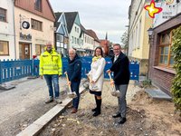Drei Männer und eine Frau stehen in einer Baugrube. Drei von ihnen haben Spaten in den Händen, als würden sie gerade in Loch buddeln.