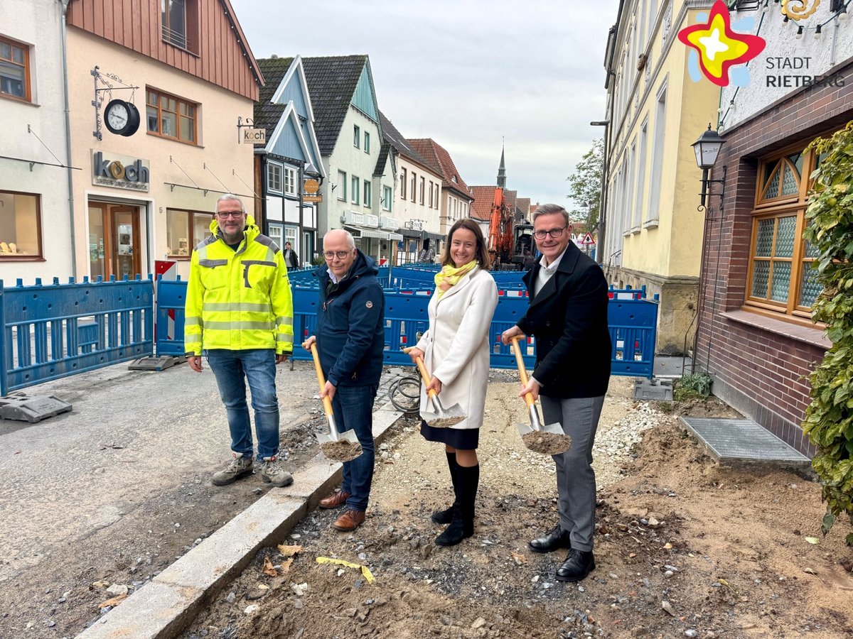 Drei Männer und eine Frau stehen in einer Baugrube. Drei von ihnen haben Spaten in den Händen, als würden sie gerade in Loch buddeln.