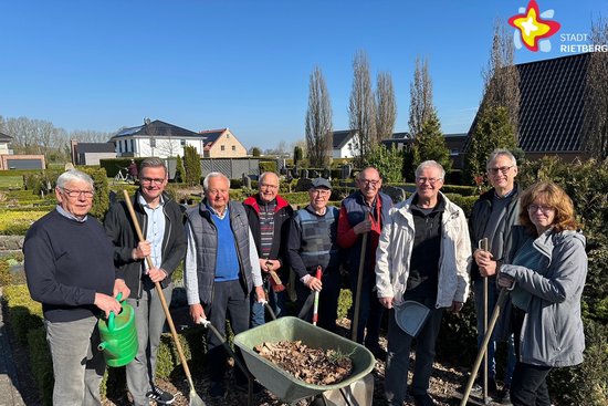 Die genannten Personen stehen nebeneinander auf dem Friedhof und schauen in die Kamera. Sie haben eine Gießkanne, eine Schubkarre und Harken in der Hand. Die Sonne scheint, der Himmerl ist blau.