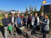 Die genannten Personen stehen nebeneinander auf dem Friedhof und schauen in die Kamera. Sie haben eine Gießkanne, eine Schubkarre und Harken in der Hand. Die Sonne scheint, der Himmerl ist blau.