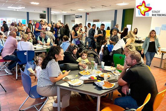 Viele Familien sitzen im Flur des Schulzentrums Rietberg an Tischen, essen und unterhalten sich.