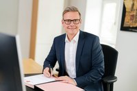 Der Bürgermeister sitzt in seinem Büro am Schreibtisch und schaut in die Kamera. Er trägt einen Anzug. Auf dem Schreibtisch liegt eine Mappe.