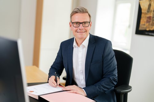 Der Bürgermeister sitzt in seinem Büro am Schreibtisch und schaut in die Kamera. Er trägt einen Anzug. Auf dem Schreibtisch liegt eine Mappe.