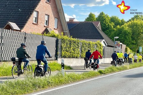 Eine Reihe Radfahrer, teils mit Helm auf dem Kopf, fährt auf einem Raweg entlang eines von einem Zaun geschützten Hauses.