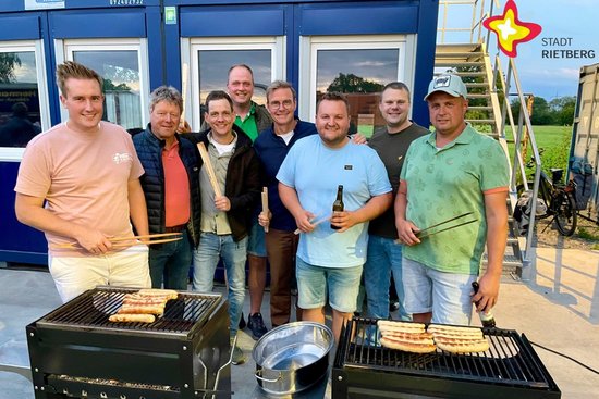 Auf zwei Grillgeräten liegen Bratwürste, dahinter stehen Teilnehmer der Tour und die genannten Personen und schauen in die Kamera.