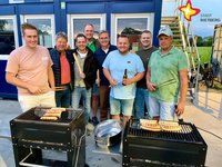 Auf zwei Grillgeräten liegen Bratwürste, dahinter stehen Teilnehmer der Tour und die genannten Personen und schauen in die Kamera.