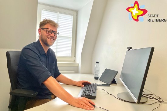 Ein Mann im schwarzen Hemd sitzt hinter einem fast leeren Schreibtisch, auf dem lediglich Tastatur und Monitor stehen.