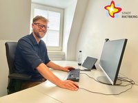Ein Mann im schwarzen Hemd sitzt hinter einem fast leeren Schreibtisch, auf dem lediglich Tastatur und Monitor stehen.