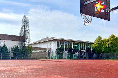 Über dem roten Kunstspielfeld schwebt im rechten Bildrand ein Basketballkorb. Im Hintergrund ist eine Sporthalle zu sehen.