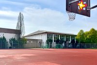 Über dem roten Kunstspielfeld schwebt im rechten Bildrand ein Basketballkorb. Im Hintergrund ist eine Sporthalle zu sehen.
