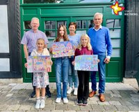 Drei Erwachsene und drei Kinder mit selbstgemalten Bildern in den Händen stehen nebeneinander vor einer grünen Tür.