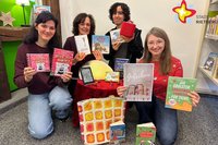 Die Mitarbeiterinnen der Stadtbibliothek knien um einen Tisch, auf dem verschiedene Bücher zu sehen sind. In der Hand halten sie Bücher und andere Medien aus der Stadtbibliothek.