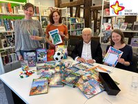 Die in der Bildzeile genannten Personen sitzen und stehen in der Stadtbibliothek an einem Tisch, auf dem Bücher ausgebreitet sind. Sie schauen in die Kamera. Im Hintergrund sind Bücherregale zu sehen.