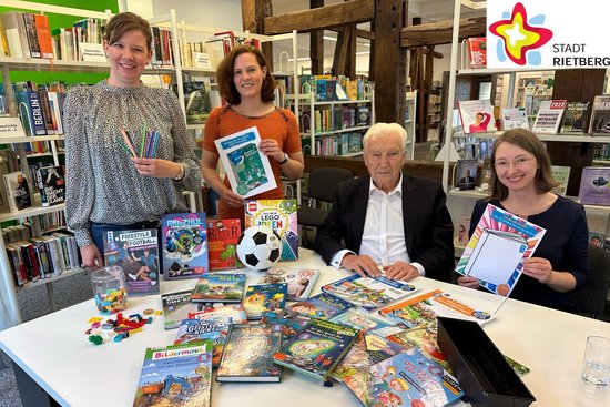 Die in der Bildzeile genannten Personen sitzen und stehen in der Stadtbibliothek an einem Tisch, auf dem Bücher ausgebreitet sind. Sie schauen in die Kamera. Im Hintergrund sind Bücherregale zu sehen.