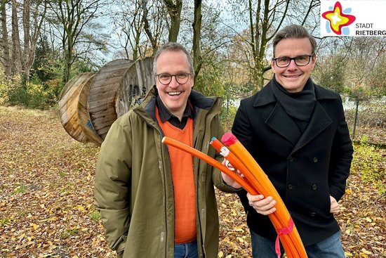Bürgermeister Andreas Sunder und der Breitbandbeauftragte der Stadt Rietberg, Rüdiger Ropinski, stehen auf einer belaubten Freifläche und halten ein dickes, mehradriges orangefarbenes Kabel in die Höhe.
