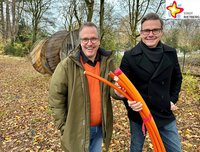 Bürgermeister Andreas Sunder und der Breitbandbeauftragte der Stadt Rietberg, Rüdiger Ropinski, stehen auf einer belaubten Freifläche und halten ein dickes, mehradriges orangefarbenes Kabel in die Höhe.