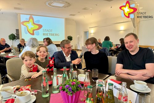 Zwei Kinder und ein Elternpaar sitzen an einem mit Blumen und Wasserflaschen gedeckten Tisch, in der Mitte kniet ein Mann mit Mikrofon in der Hand und hält es der Mutter vor den Mund.