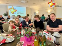 Zwei Kinder und ein Elternpaar sitzen an einem mit Blumen und Wasserflaschen gedeckten Tisch, in der Mitte kniet ein Mann mit Mikrofon in der Hand und hält es der Mutter vor den Mund.