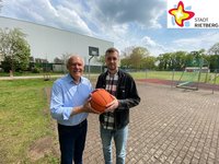 Zwei Männer stehen unter freiem Himmel auf dem Basketballplatz und halten einen Ball in der Hand. Im Hintergrund ist einer der Basketballkörbe zu sehen. Die Sonne scheint.