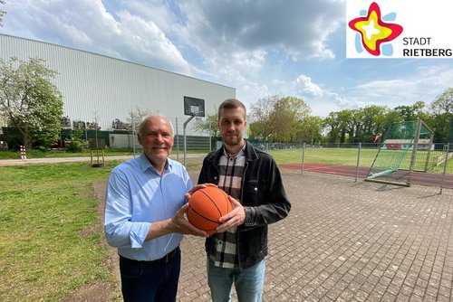 Zwei Männer stehen unter freiem Himmel auf dem Basketballplatz und halten einen Ball in der Hand. Im Hintergrund ist einer der Basketballkörbe zu sehen. Die Sonne scheint.