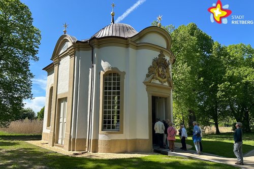 Einige Menschen stehen vor der Tür einer kleinen, weißen Kapelle. Der Himmel strahlt blau.
