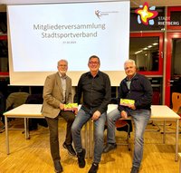 Dre Männer sitzen lächelnd nebeneinander auf einem Tisch. Im Hintergrund ist auf einer Leinwand der Schriftzug "Mitgliederversammlung Stadtsportverband" zu lesen.