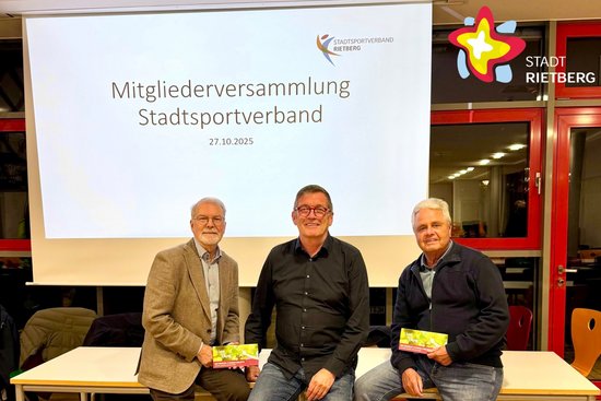 Dre Männer sitzen lächelnd nebeneinander auf einem Tisch. Im Hintergrund ist auf einer Leinwand der Schriftzug "Mitgliederversammlung Stadtsportverband" zu lesen.
