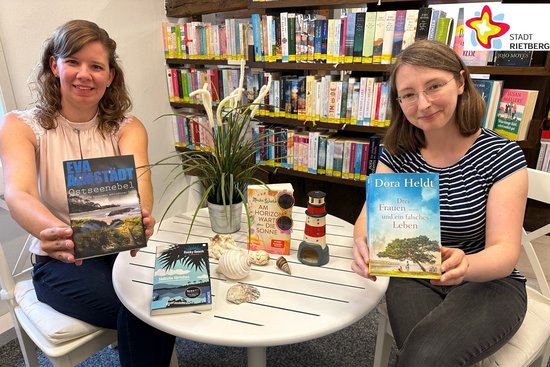 Jennifer Bader und Julia Bröckling sitzen an einem runden Tisch und halten Bücher in der Hand. Auf dem Tisch liegen zwei weitere Bücher sowie verschiedene Muscheln und ein Modell von einem Leuchtturm. Im Hintergrund ist ein Bücheregal zu sehen.