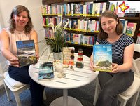 Jennifer Bader und Julia Bröckling sitzen an einem runden Tisch und halten Bücher in der Hand. Auf dem Tisch liegen zwei weitere Bücher sowie verschiedene Muscheln und ein Modell von einem Leuchtturm. Im Hintergrund ist ein Bücheregal zu sehen.