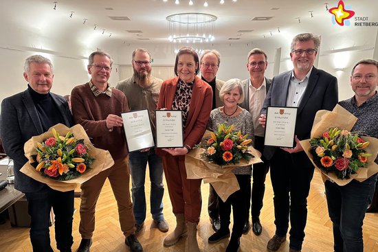 Die genannten Personen stehen nebeneinander im Ratssaal. Drei halten Urkunden in den Händen, weitere drei Personen Blumensträuße. Alle schauen in die Kamera und lächeln.