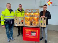 Die genannten Personen schauen in die Kamera. Sie stehen und präsentieren das Rauchdemohaus, das so groß ist wie ein Puppenhaus.
